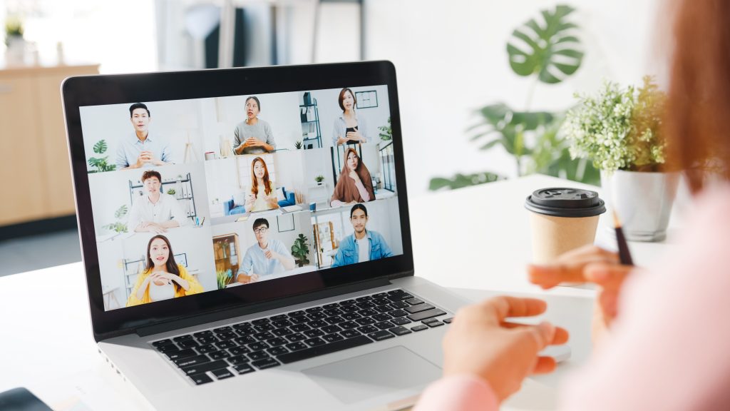 Person sitzt an Schreibtisch vor Laptop mit einer Videokonferenz mit mehreren Personen auf dem Bildschirm.