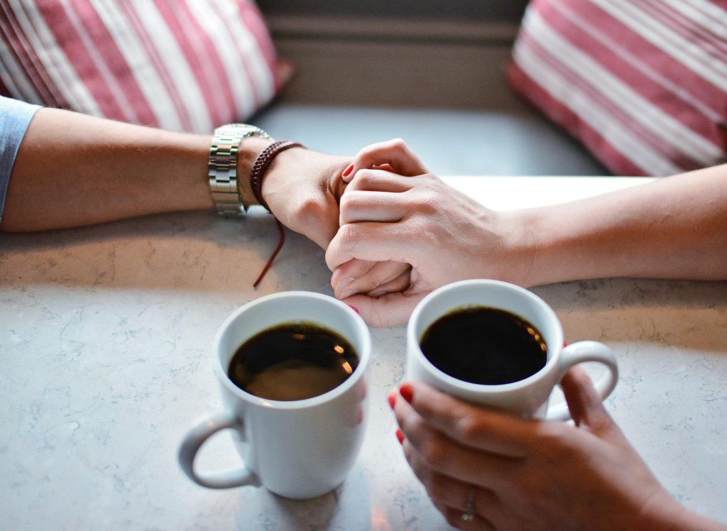 Zwei Personen verschränken ihre Finger ineinander. Neben ihnen stehen zwei Tassen Kaffee auf dem Tisch.